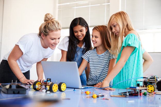 Drei Mädchen programmieren ein Roboterfahrzeug mit einer Lehrperson am Laptop.
